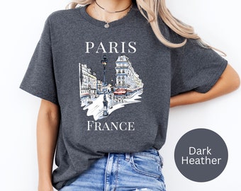 May include: Dark heather grey t-shirt with a white graphic of a street scene in Paris, France. The graphic includes buildings, a street lamp, and people walking. The text "PARIS" and "FRANCE" is printed in white on the shirt.