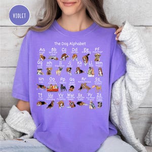 May include: A purple t-shirt with a graphic of a dog alphabet. Each letter of the alphabet is paired with a dog breed and a word that starts with that letter. For example, 'A' is for 'Angel' and 'B' is for 'Belly Rub Lover'.