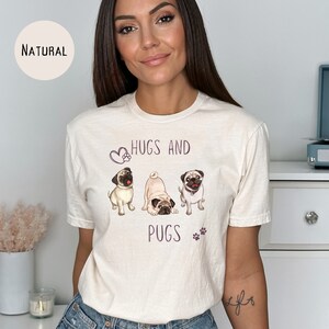 Puede incluir: Una camiseta blanca con un gr&aacute;fico de tres carlinos y el texto "Hugs and Pugs" en morado. Los carlinos est&aacute;n en una pose juguetona, con un carlino mirando a la c&aacute;mara.