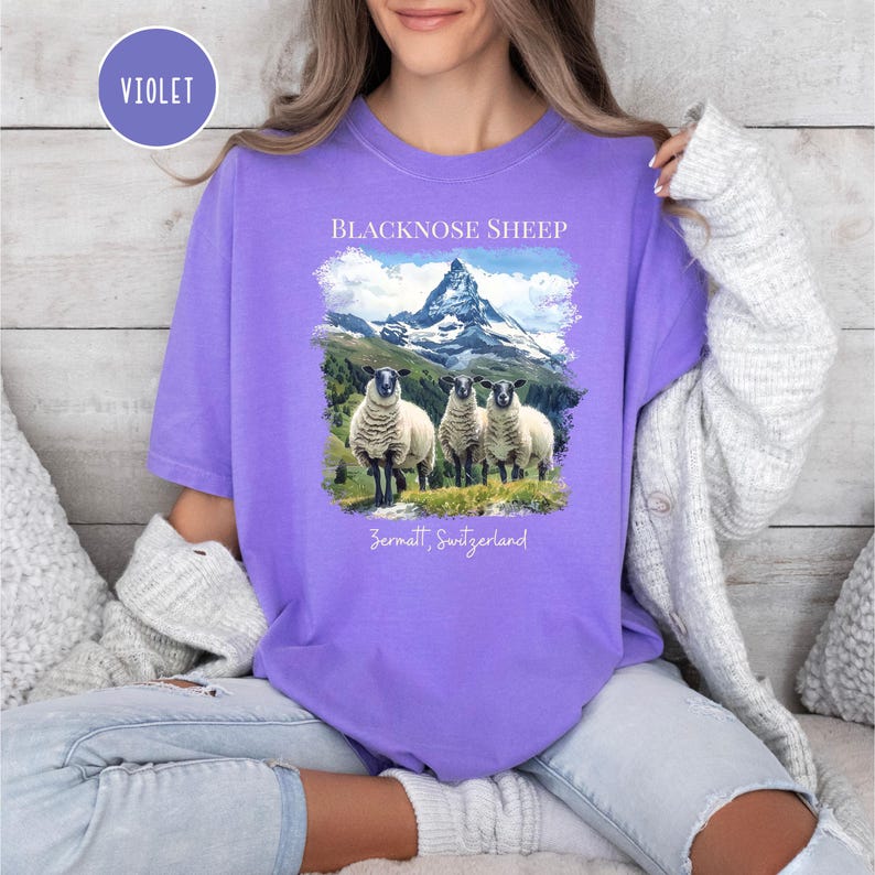 K&ouml;nnte beinhalten: Ein violettes T-Shirt mit einer Grafik von drei Schwarznasenschafen vor einer Berglandschaft. Der Text "BLACKNOSE SHEEP" und "Zermatt, Switzerland" sind aufgedruckt. Das Shirt ist aus weichem Material.