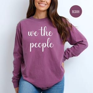 Puede incluir: Una camiseta de manga larga de color morado con el texto "we the people" impreso en blanco en la parte delantera.