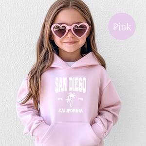 May include: A light pink hoodie with the text "SAN DIEGO CALIFORNIA" in white. The hoodie features a palm tree graphic and the text "EST. 1769". The person in the image is wearing pink heart-shaped sunglasses.