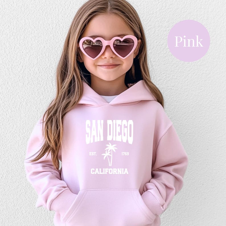 May include: A light pink hoodie with the text "SAN DIEGO CALIFORNIA" in white. The hoodie features a palm tree graphic and the text "EST. 1769". The person in the image is wearing pink heart-shaped sunglasses.