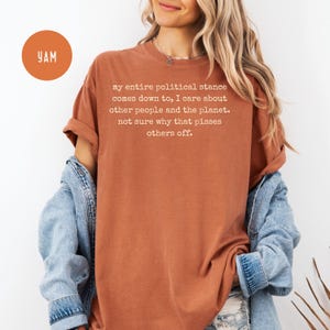 May include: Rust-colored t-shirt with the text "my entire political stance comes down to, I care about other people and the planet. not sure why that pisses others off." A round orange sticker with "YAM" is in the upper left.