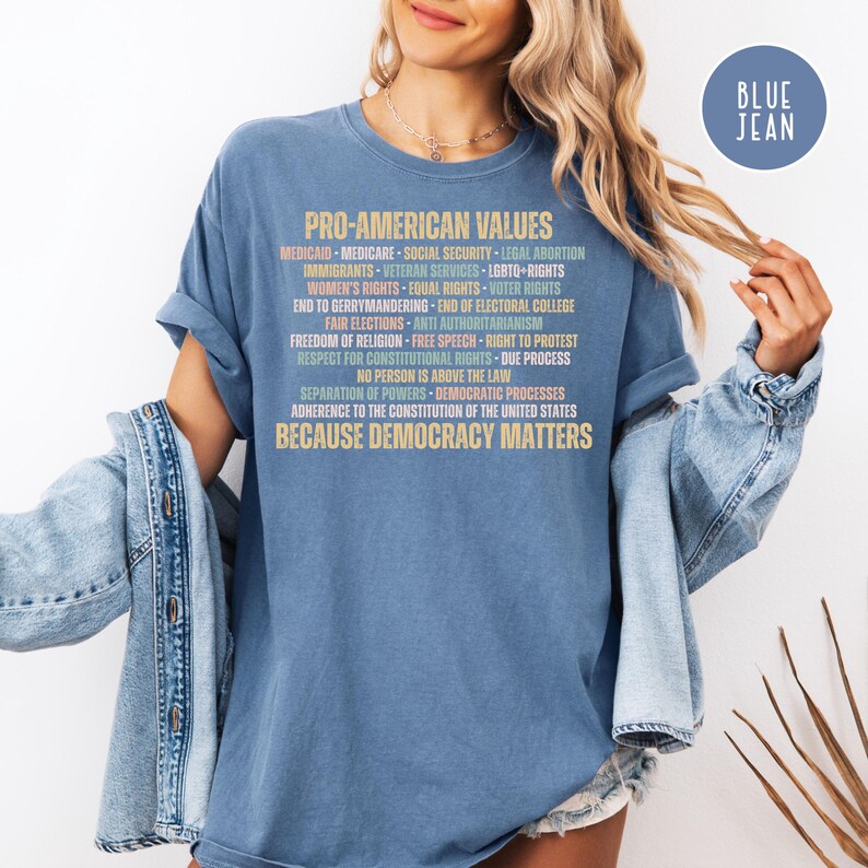 Pode incluir: Uma camiseta azul com o texto "PRO-AMERICAN VALUES" em formato empilhado. O texto lista v&aacute;rios valores e princ&iacute;pios, incluindo "Medicaid", "Seguran&ccedil;a Social" e "Democracy Matters". Um logotipo "Blue Jean" est&aacute; no canto superior direito.