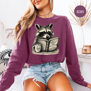 May include: A berry-colored long-sleeve sweatshirt featuring a graphic of a raccoon wearing glasses, reading a book titled How to Protect Our Capitol from a Hostile Takeover and Citizens from a Corrupt President, with a mug labeled Is It 2028 Yet.