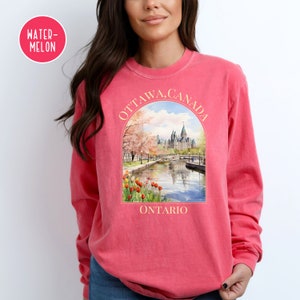 May include: A pink long-sleeved t-shirt with a watercolor illustration of the Parliament Buildings in Ottawa, Canada. The text "OTTAWA, CANADA" is written in an arch above the image, and "ONTARIO" is written below the image.