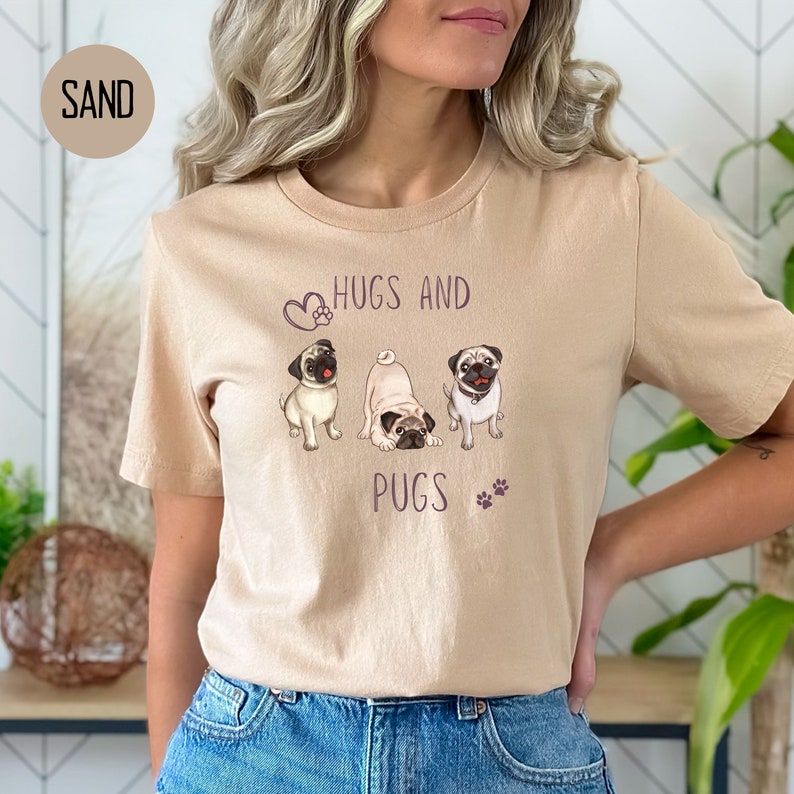 Puede incluir: Una camiseta de color arena con un gr&aacute;fico de tres carlinos y el texto "Hugs and Pugs" en morado.