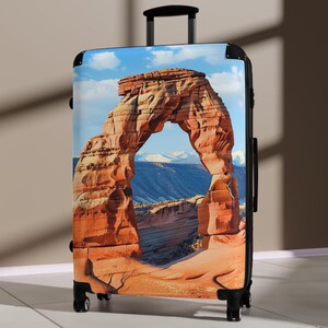 May include: A black suitcase with a printed image of a large sandstone archway in a desert landscape. The archway is a natural rock formation with a blue sky and clouds in the background.