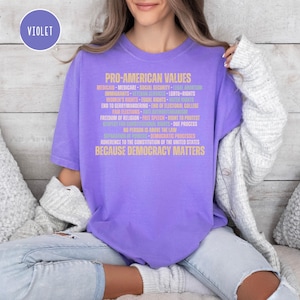 Pode incluir: Uma camiseta violeta com o texto dourado "PRO-AMERICAN VALUES". O texto lista v&aacute;rios valores pol&iacute;ticos e sociais, incluindo "Medicaid", "Seguran&ccedil;a Social" e "Direitos LGBTQ+". A frase "BECAUSE DEMOCRACY MATTERS" est&aacute; na parte inferior.