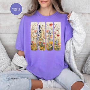May include: A purple t-shirt with a floral print design. The design features five vertical panels of watercolor flowers in shades of pink, yellow, blue, and green.