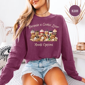 May include: A berry-colored long-sleeve sweatshirt featuring a graphic print of various coffee cups and assorted cookies. The text reads, "Because a Cookie Lover Needs Options." The design includes detailed illustrations of beverages and baked goods.