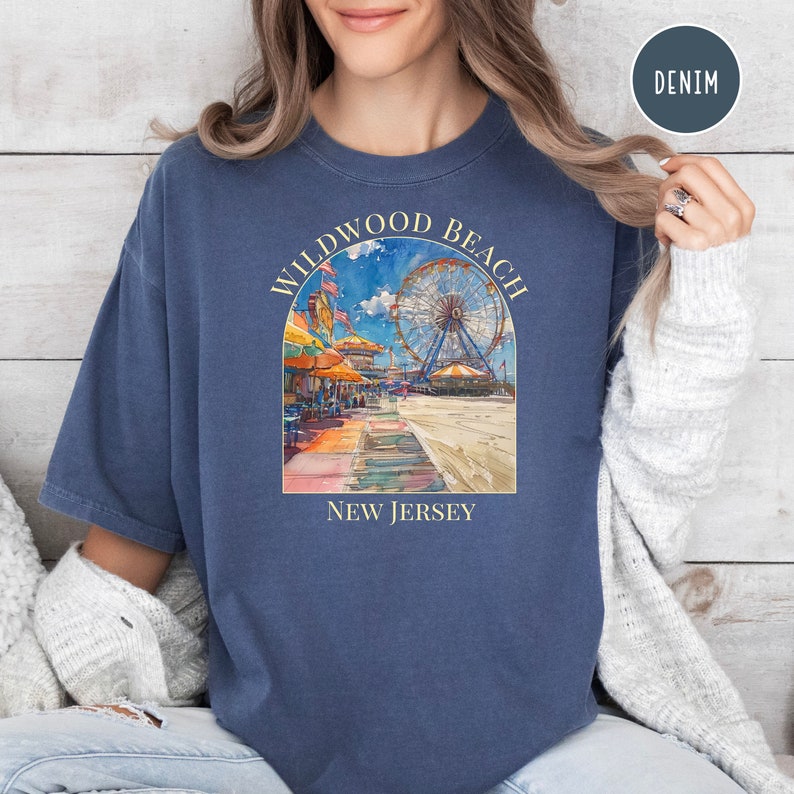 May include: A navy blue t-shirt with a watercolor illustration of the Wildwood Beach Ferris wheel and boardwalk. The text "Wildwood Beach" is in a yellow arch above the illustration and "New Jersey" is below.