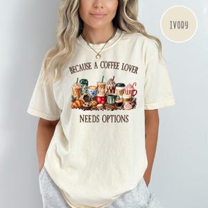 May include: An ivory short-sleeve t-shirt featuring a graphic print of various coffee drinks, pastries, and coffee beans. The text reads "BECAUSE A COFFEE LOVER NEEDS OPTIONS" in brown lettering. The shirt has a crew neckline and a relaxed fit.