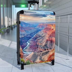 May include: A black suitcase with a colorful image of the Grand Canyon on the front. The image shows a river winding through the canyon, with layered rock formations in shades of red, orange, brown, and purple.