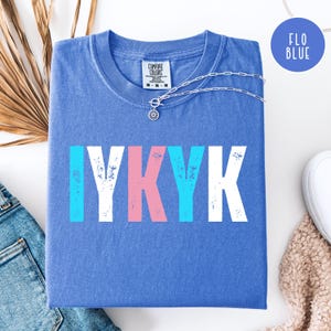 Puede incluir: Una camiseta azul Comfort Colors con el texto "IYKYK" en blanco, rosa y azul. Un collar plateado con un colgante de sol descansa sobre la camiseta. La camiseta está doblada sobre una superficie blanca, con unos vaqueros y una bufanda beige visibles.