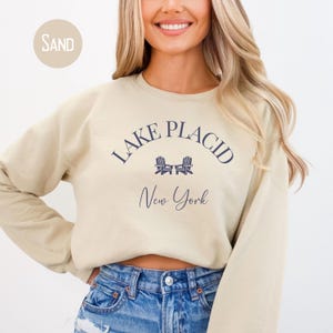 May include: Sand-colored sweatshirt with the text "Lake Placid New York" in navy blue. The design includes two chairs. The sweatshirt is a crew neck style.