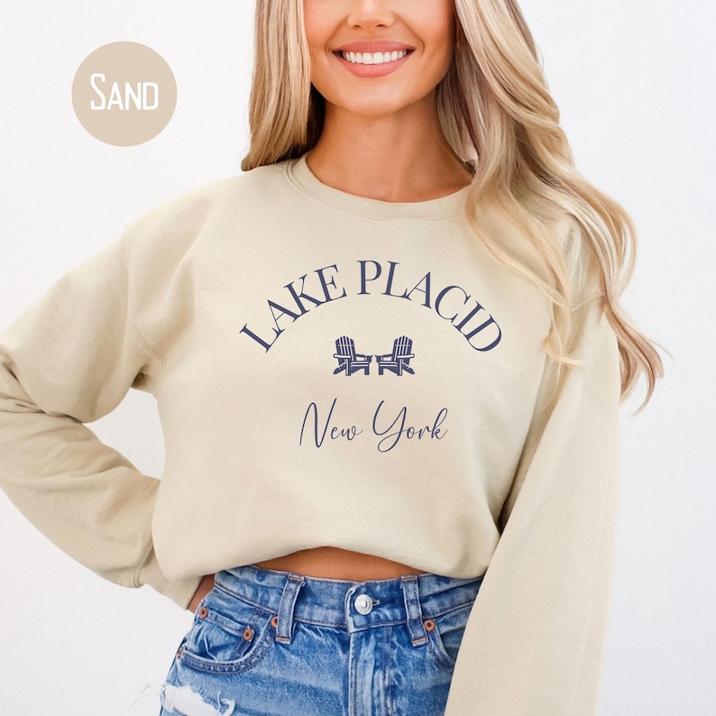 May include: Sand-colored sweatshirt with the text "Lake Placid New York" in navy blue. The design includes two chairs. The sweatshirt is a crew neck style.