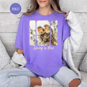 May include: A purple t-shirt with a graphic of three birds in a nest with flowers and the text "Spring is Here".