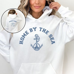 May include: White hooded sweatshirt with a blue anchor and the text "Home by the Sea" printed on the front. The back of the sweatshirt has the text "Hamptons New York" with a sailboat graphic.
