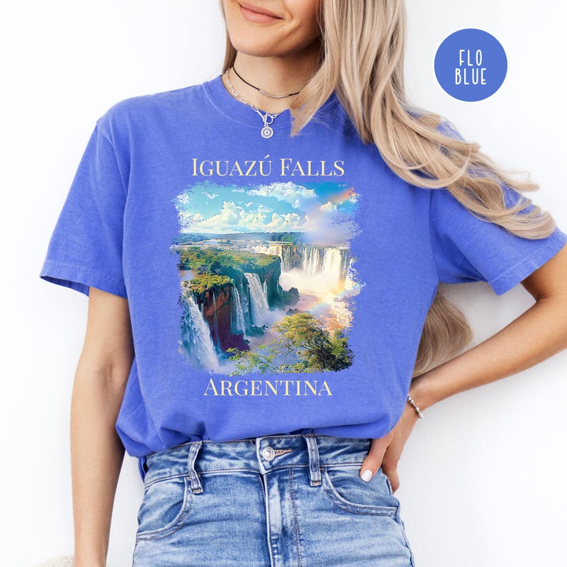 Pu&ograve; includere: T-shirt blu con un'immagine delle cascate dell'Iguaz&uacute; in Argentina. Il design include il testo "IGUAZ&Uacute; FALLS" e "ARGENTINA". La maglietta ha un girocollo e maniche corte.