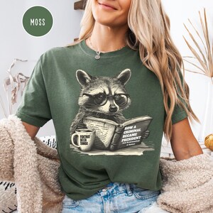 May include: Moss green t-shirt featuring a raccoon wearing glasses and reading a book titled "How a Criminal Became President." A coffee mug with "Foxtrot Delta Tango" is on the left. The shirt has a relaxed fit.