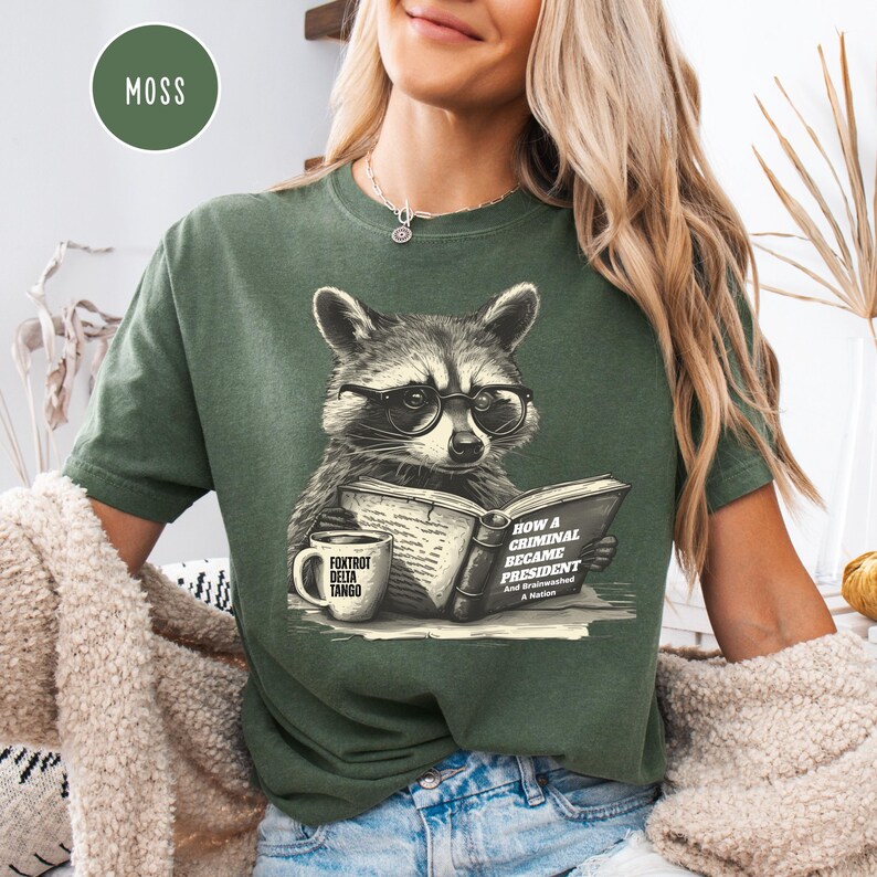 May include: Moss green t-shirt featuring a raccoon wearing glasses and reading a book titled "How a Criminal Became President." A coffee mug with "Foxtrot Delta Tango" is on the left. The shirt has a relaxed fit.