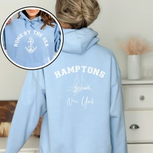 May include: Light blue hooded sweatshirt with white text and graphic on the back. The text reads "HAMPTONS New York" and the graphic is a sailboat. The front of the sweatshirt has white text that reads "HOME BY THE SEA" with an anchor graphic.