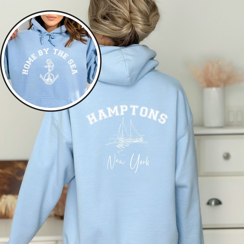 May include: Light blue hooded sweatshirt with white text and graphic on the back. The text reads "HAMPTONS New York" and the graphic is a sailboat. The front of the sweatshirt has white text that reads "HOME BY THE SEA" with an anchor graphic.