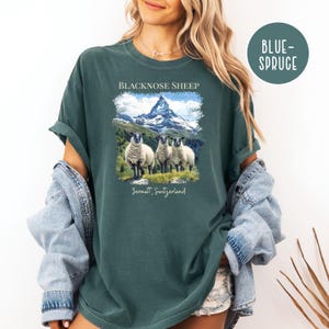 K&ouml;nnte beinhalten: Blau-Fichte farbenes T-Shirt mit einer Grafik von drei Schwarznasenschafen vor einer Bergkette. Der Text "BLACKNOSE SHEEP" und "Zermatt, Switzerland" sind auf dem Shirt aufgedruckt.