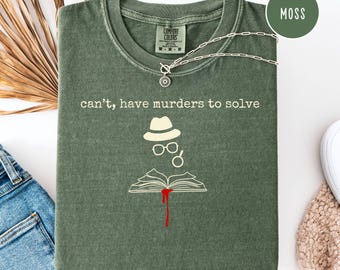 Peut inclure: T-shirt vert mousse avec l'inscription "can't, have murders to solve" et un dessin de détective. Le dessin comprend un chapeau, des lunettes et un livre ouvert avec une goutte de sang. Un collier argenté avec un pendentif soleil est sur le t-shirt.