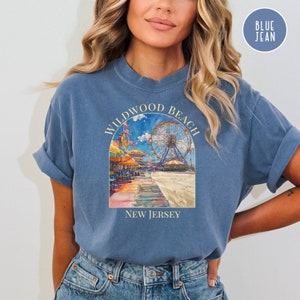 May include: A blue jean t-shirt with a graphic of a Ferris wheel and boardwalk scene. The text "Wildwood Beach New Jersey" is printed on the shirt.