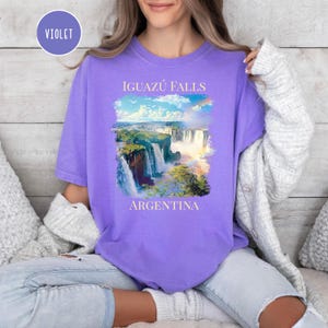 Pu&ograve; includere: T-shirt viola con un'immagine delle cascate dell'Iguaz&uacute; in Argentina. Il design include il testo "IGUAZ&Uacute; FALLS" sopra l'immagine e "ARGENTINA" sotto. La maglietta &egrave; di colore viola unito.
