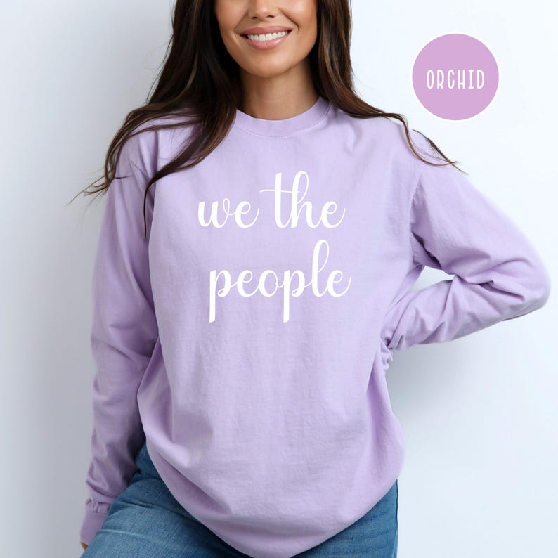 Puede incluir: Una camiseta de manga larga de color lila claro con el texto "we the people" impreso en blanco en la parte delantera.