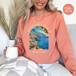 K&ouml;nnte beinhalten: Ein korallenfarbenes Sweatshirt mit einem Grafikdruck von Positano, Italien, an der Amalfik&uuml;ste. Die Grafik zeigt eine farbenfrohe Stadtlandschaft mit einem blauen Meer und einem Zitronenbaum.