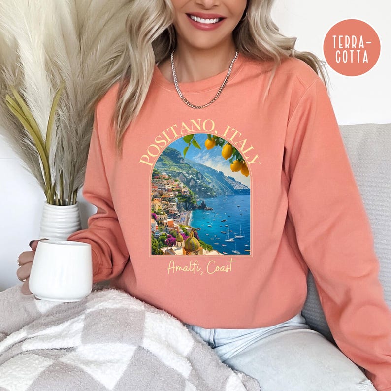 K&ouml;nnte beinhalten: Ein korallenfarbenes Sweatshirt mit einem Grafikdruck von Positano, Italien, an der Amalfik&uuml;ste. Die Grafik zeigt eine farbenfrohe Stadtlandschaft mit einem blauen Meer und einem Zitronenbaum.