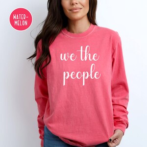 Puede incluir: Una camiseta rosa de manga larga con el texto "we the people" en letras blancas.