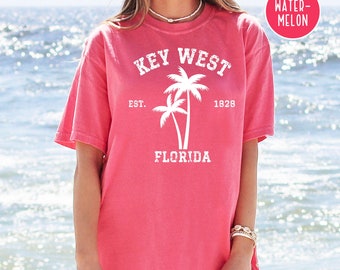 May include: A pink t-shirt with the text "KEY WEST EST. 1928 FLORIDA" and a palm tree graphic. The shirt is a relaxed fit and has a crew neck.