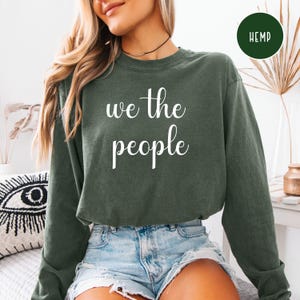 Puede incluir: Una camiseta verde de manga larga con el texto "we the people" en letras blancas.