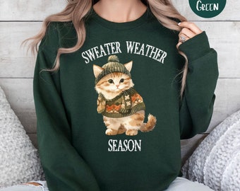 以下が含まれることがあります： 「Sweater Weather Season」という文字と、セーターと帽子をかぶった漫画の猫が描かれた、森の緑色のスウェットシャツです。