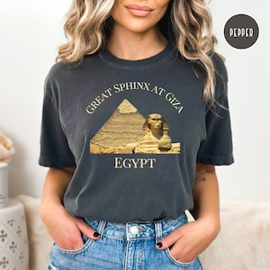 May include: A dark gray t-shirt with a graphic of the Great Sphinx at Giza in Egypt. The graphic is in gold and features the Sphinx and the Great Pyramid of Giza.