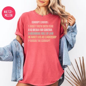May include: A watermelon-colored t-shirt with the text "CORRUPT LEADERS: TARGET TRUTH WITH FEAR, RIG MEDIA FOR CONTROL, UNDERMINE RULE OF LAW, MARKET LIES AS LEADERSHIP, PARDON THE CORRUPT." The shirt is a relaxed fit.