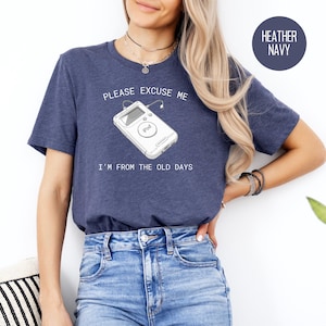 Pode incluir: Uma camiseta azul marinho desbotada com um gráfico branco de um iPod e o texto "Por favor, desculpe-me, eu sou da velha guarda."