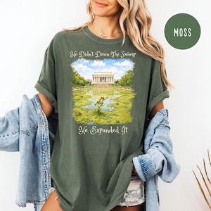 May include: A moss green short-sleeve t-shirt featuring a graphic print of the Lincoln Memorial surrounded by a swamp with a frog in the foreground. The text reads, He Didn't Drain The Swamp, He Expanded It. The shirt is worn with a light blue denim jacket.