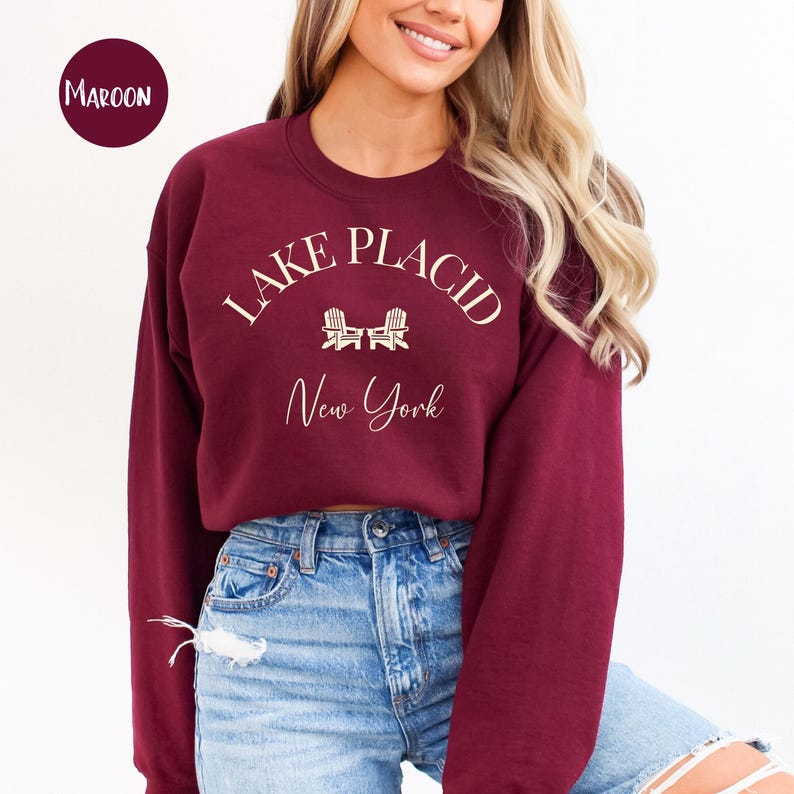 May include: Maroon crewneck sweatshirt with the text "Lake Placid New York" in a white script font. The design includes two small white Adirondack chairs. The word "Maroon" is in a circle in the upper left corner.