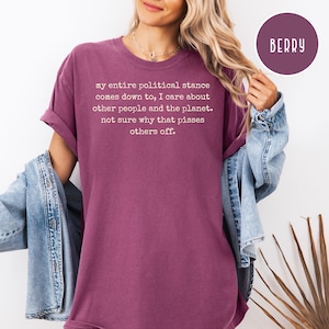 May include: Berry-colored t-shirt with rolled-up sleeves. The shirt features white text: "my entire political stance comes down to, I care about other people and the planet. not sure why that pisses others off." A circular "BERRY" label is in the upper right.