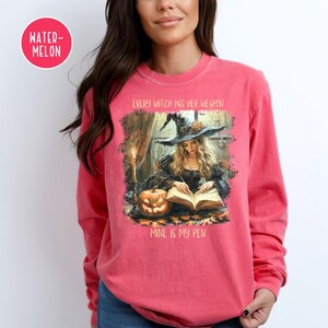 May include: A coral-colored long-sleeve shirt featuring a witch-themed graphic. The graphic depicts a witch reading a book with a pumpkin and candle. Text on the shirt reads "EVERY WITCH HAS HER WEAPON, MINE IS MY PEN."