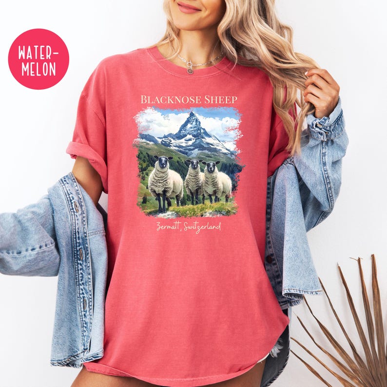 K&ouml;nnte beinhalten: Rotes T-Shirt mit einer Grafik von drei Schwarznasenschafen vor einer Bergkette. Der Text "BLACKNOSE SHEEP" und "Zermatt, Switzerland" sind oberhalb und unterhalb des Bildes aufgedruckt. Das T-Shirt hat kurze &Auml;rmel.