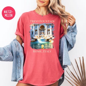 May include: A coral-coloured t-shirt with a graphic of the Trevi Fountain in Rome, Italy. The design includes the text "TREVI FOUNTAIN" above the image and "ROME, ITALY" below. The shirt has rolled-up sleeves.