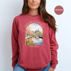 May include: A crimson long-sleeve t-shirt with a graphic print of the Parliament Building in Ottawa, Canada. The graphic features a watercolor style illustration of the building with cherry blossoms and a river in the foreground. The text "OTTAWA, CANADA" is printed in a cream color above the graphic, and the text "ONTARIO" is printed in a cream color below the graphic.